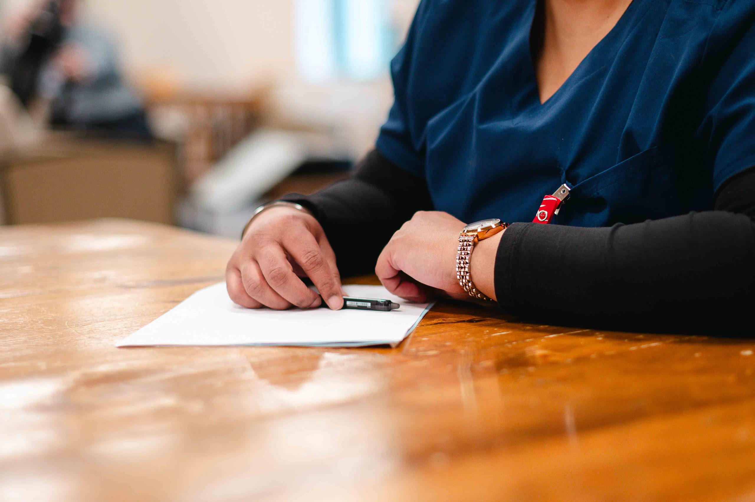 A woman in scrubs leaning on her forearms on a table and holding a pen and paper.