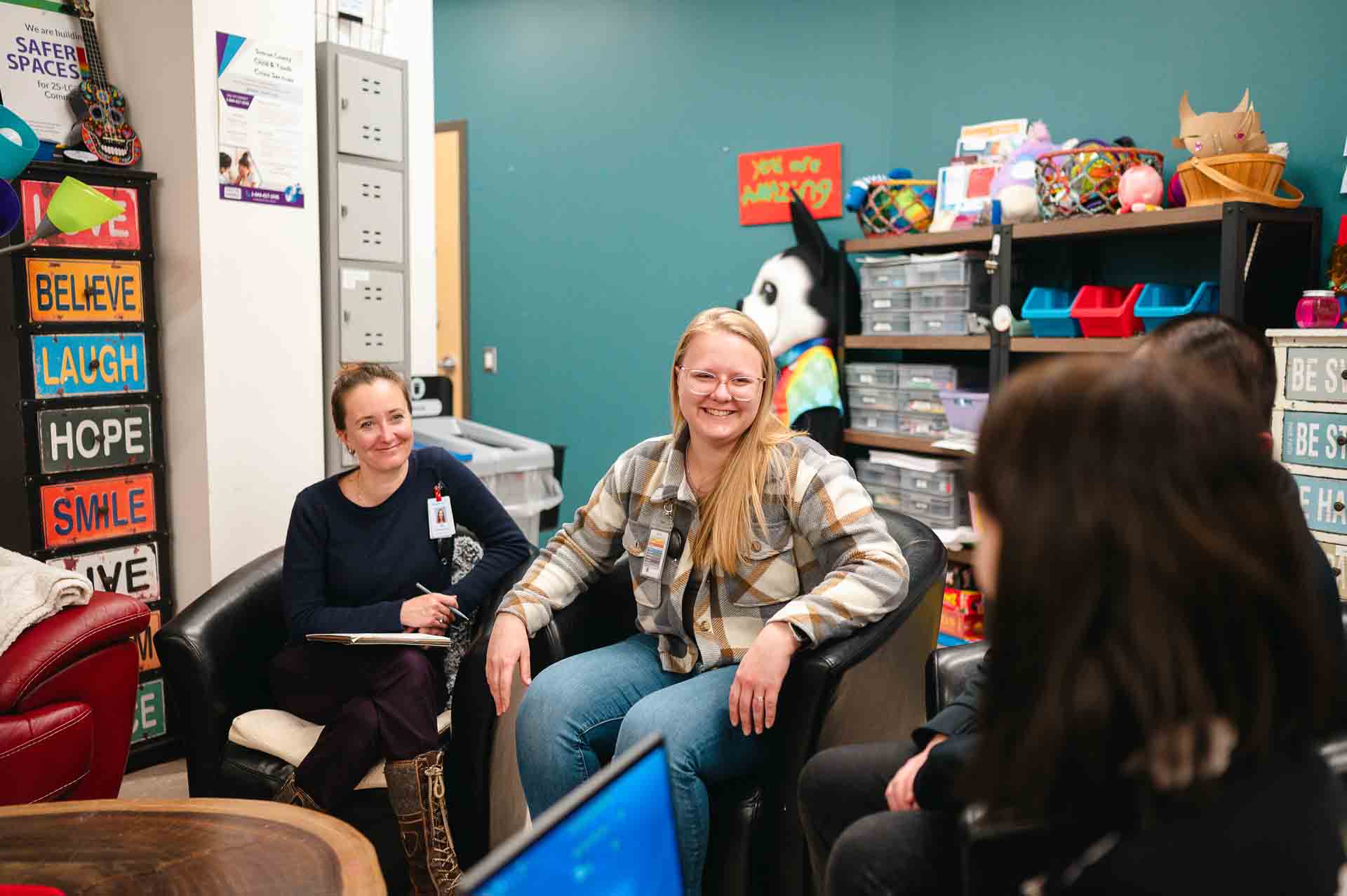Three women sit in a room laughing together with laptops and notepads around.