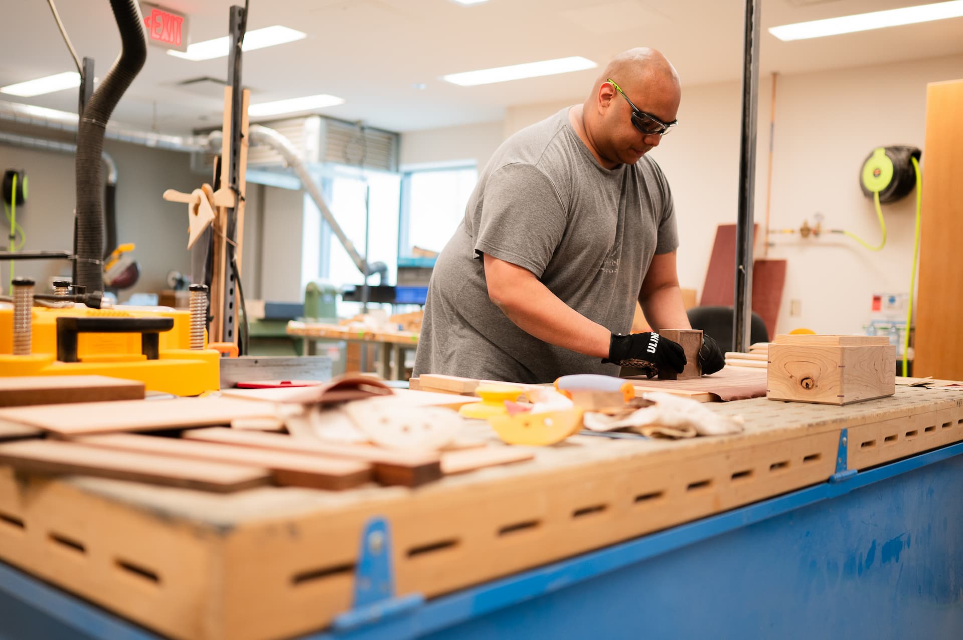 A man standing at a workbench in a workshop wearing goggles and gloves.