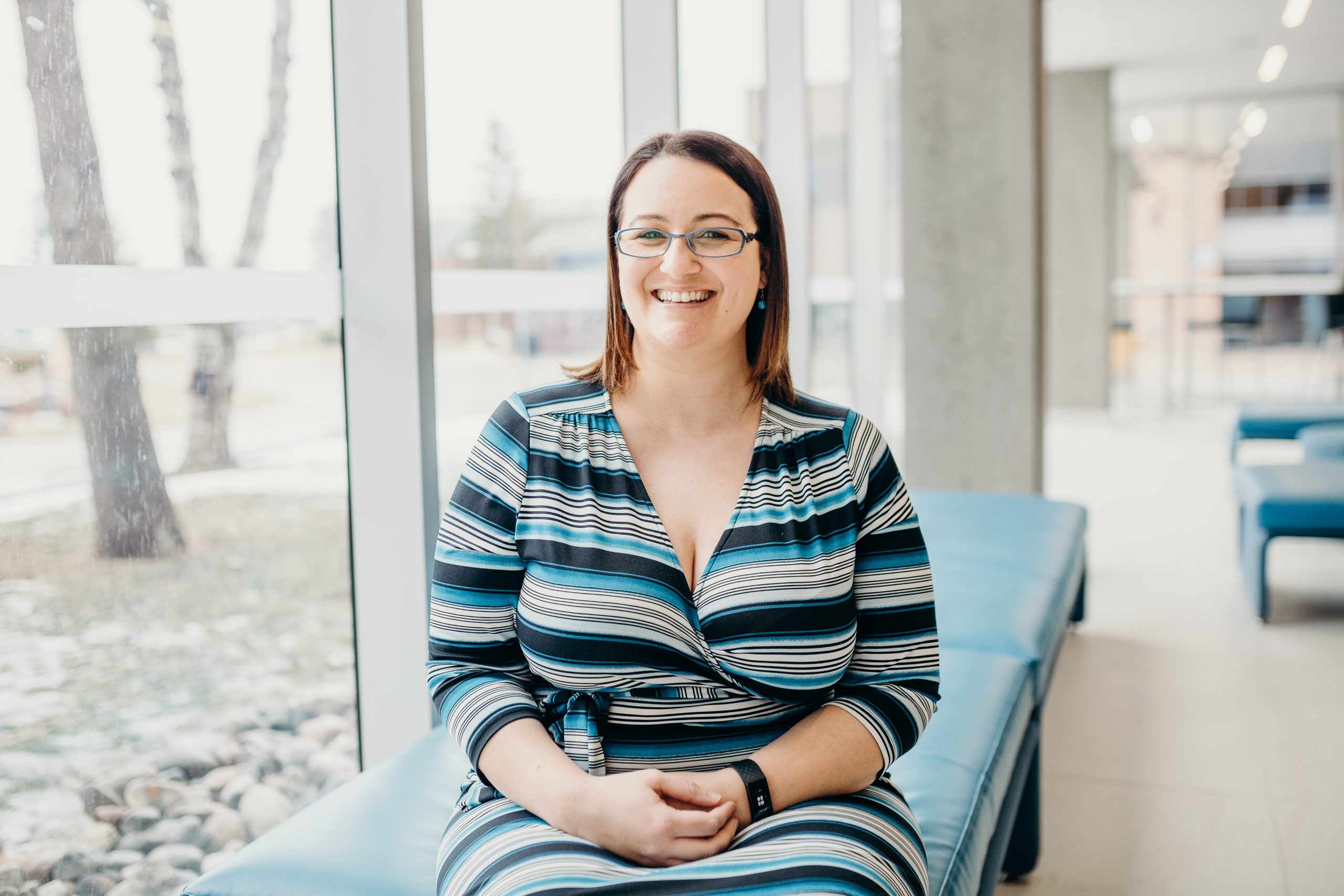 A headshot of Laura Ball sitting on a bench in front of a window overlooking a forest.