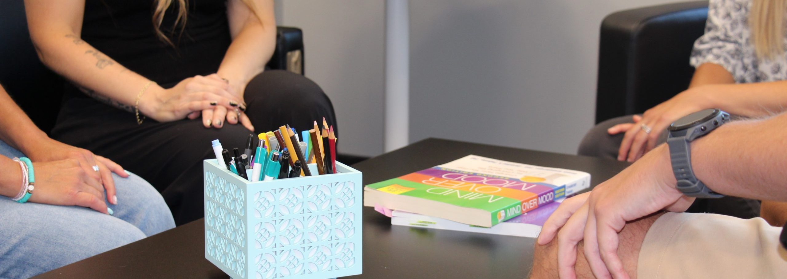 A group of four people sitting around a coffee table that has a box of pencil crayons and cognitive behavioural therapy books on top of it.