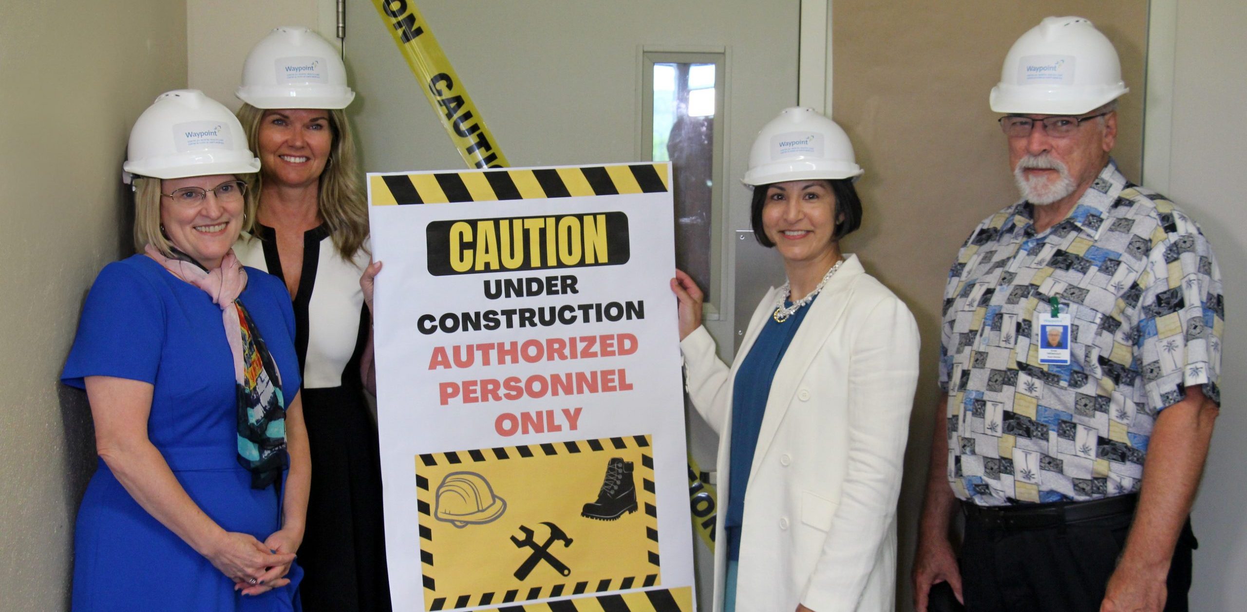 Dr. Sunderji, Minister Jones, Minister Dunlop and Ernie Vallancourt, wearing hardhats and holding a sign that reads "Caution: Under Construction."
